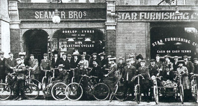 Seamer Brothers bicycle shop
Advertisements in window show "Dunlop Tyres" and "Ride electric cycles". Also shows the adjacent front of the Star Furnishing Company. The group of cyclists includes one man on a bicycle with a woman in a basket-chair trailer, and another with a woman in a sidecar.
Keywords: bicycles;shops