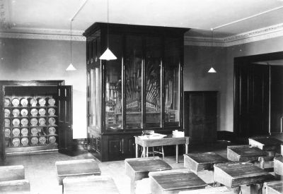 Orchestrion, by Imhof and Mukle
This mechanical musical instrument in a wooden cabinet appears to be made by Imhof & Mukle (the name plate is just distinguishable). The firm was established in 1874 in the Black Forest region of Germany, and they later had a showroom in New Oxford Street, London. The music was recorded as pins on large cylinders, and 28 of these can be seen in the adjoining cabinet. Such instruments were produced towards the end of the 19th century, preceding those which used punched paper rolls and before phonographs and gramophones became available.
This appears to be in a schoolroom. The picture came from an Enfield photographer, so there is a good chance that it was in Enfield. Does anyone remember this instrument, or recognize the room? The distinctive cornice might still exist.
Keywords: 19th century;music;musical instruments;schools