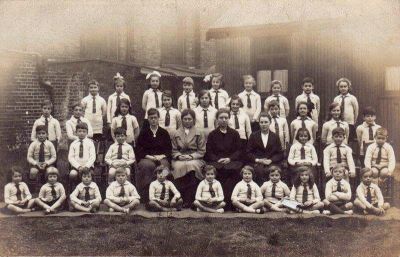 Group of children and teachers from an unidentifed primary school
This picture was probably taken in the 1930s by Frederick Roberts of  Manor Road, Enfield, a freelance photographer for the Enfield Gazette etc., who is known to have worked in the period 1917–1949. Most of his photographs are not far from Manor Road. This school picture might be St Andrews, St Georges, St Michaels, Chase Side or Lavender. The windows in the background on the left of the picture look like a church, but it is made with bricks; St Michaels church, Gordon Hill, is stone faced.  On the right is a single story corrugated iron building.  Stephen Sellick is fairly sure that it is not the old St Andrews School, Sydney Road.  Can anyone identify it?
Keywords: 1930s;schools;children